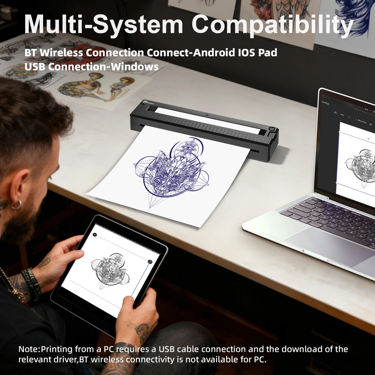 Person using a tattoo design app on a tablet with a printer and laptop on a desk.